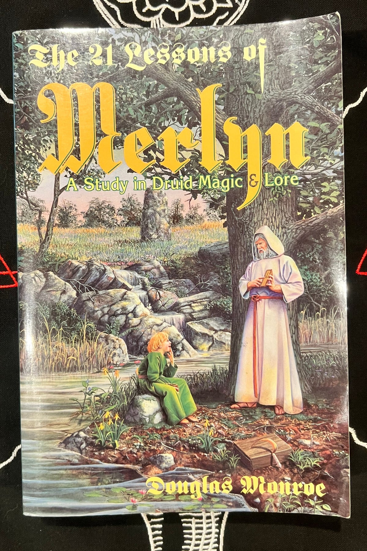 The 21 Lessons of Merlyn paperback book by Douglas Montive, featuring a detailed cover illustration of a forest scene with figures near a stream, displayed on a black background with decorative white line drawings.
