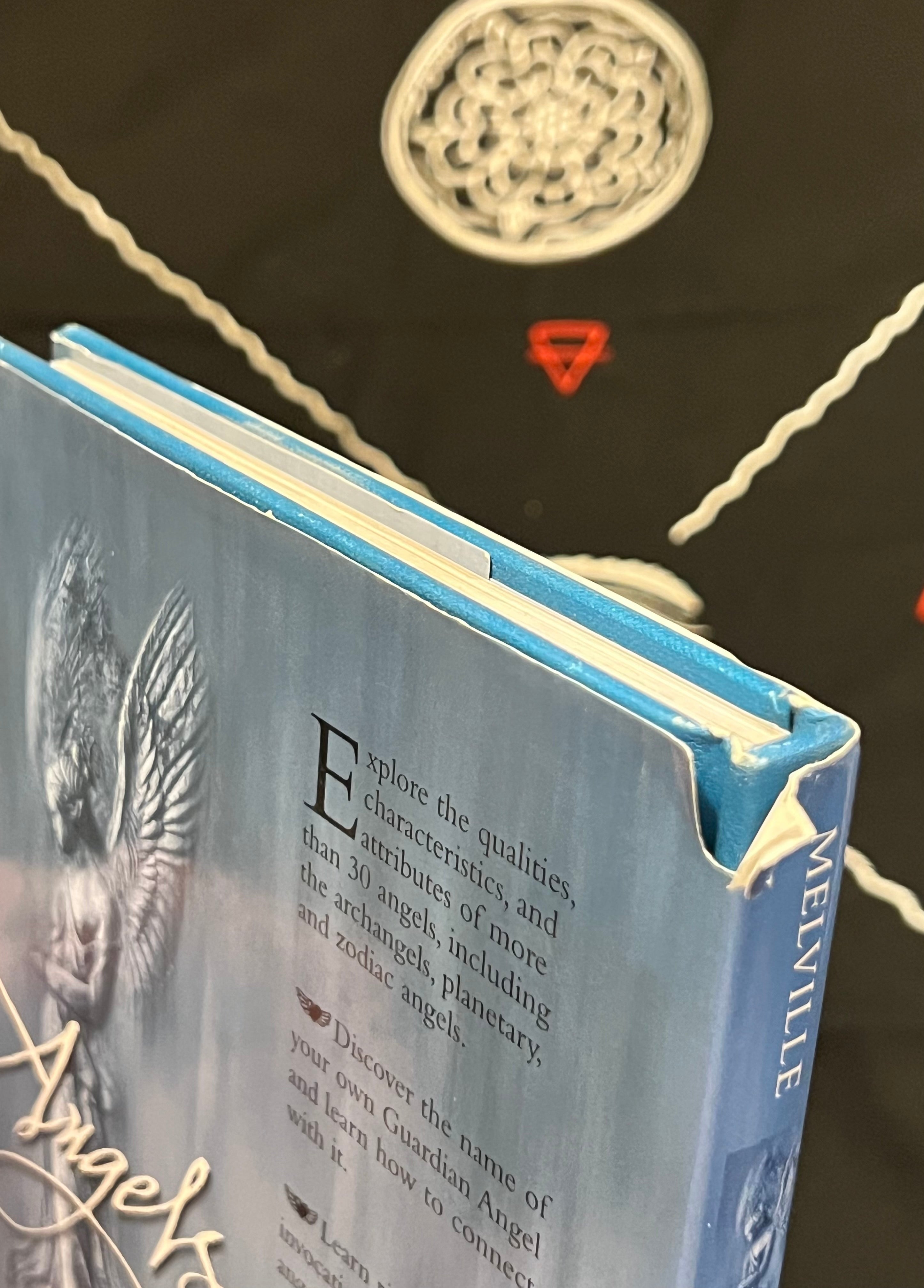 A close-up of a hardcover book titled 'Angels' by Melville, featuring a light blue cover with an angel illustration and text, set against a dark background with white rope and a circular metal ornament.