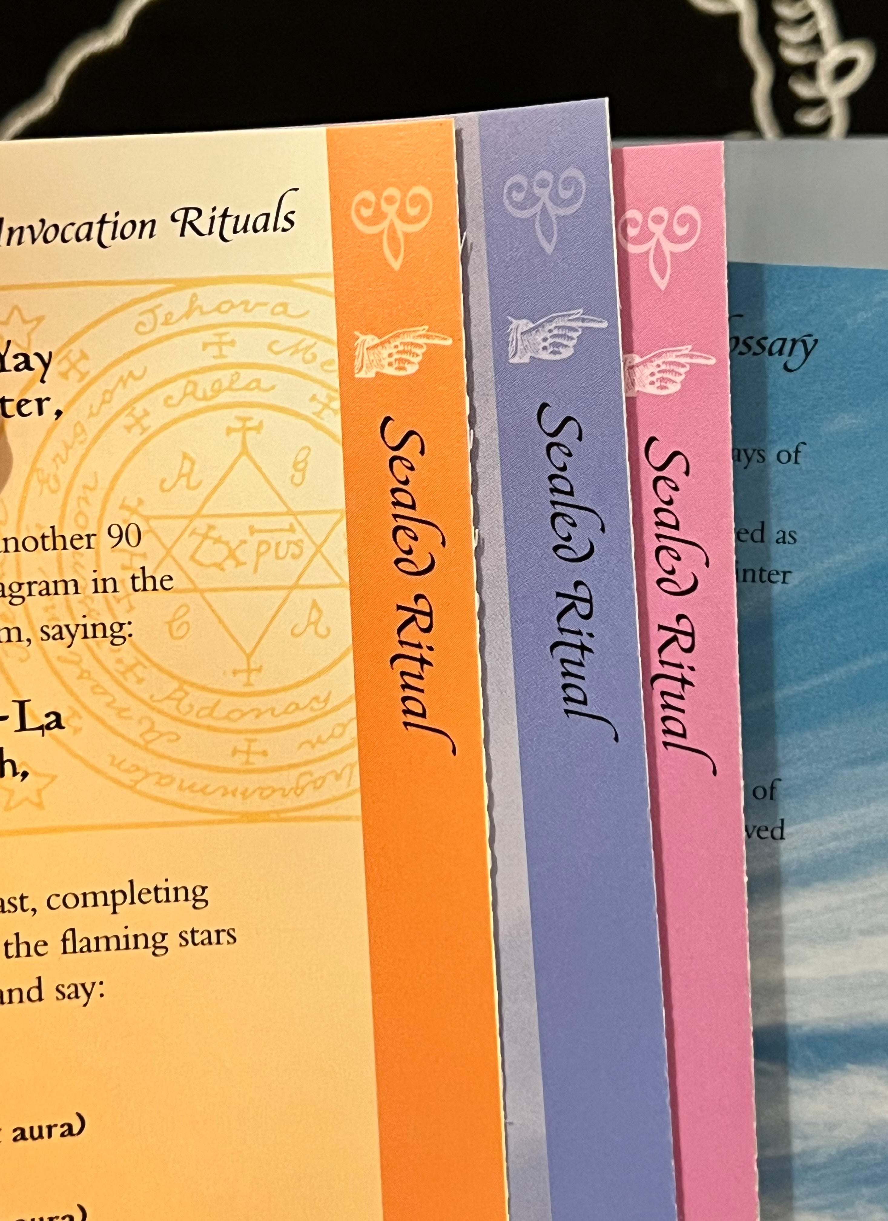 A close-up of several ritual booklets or cards standing upright, showing their colorful spines and partial covers. Visible titles include 'Sealed Ritual' in orange, lavender, and pink, alongside a cream cover with a yellow occult-style symbol and a blue cover edge.
