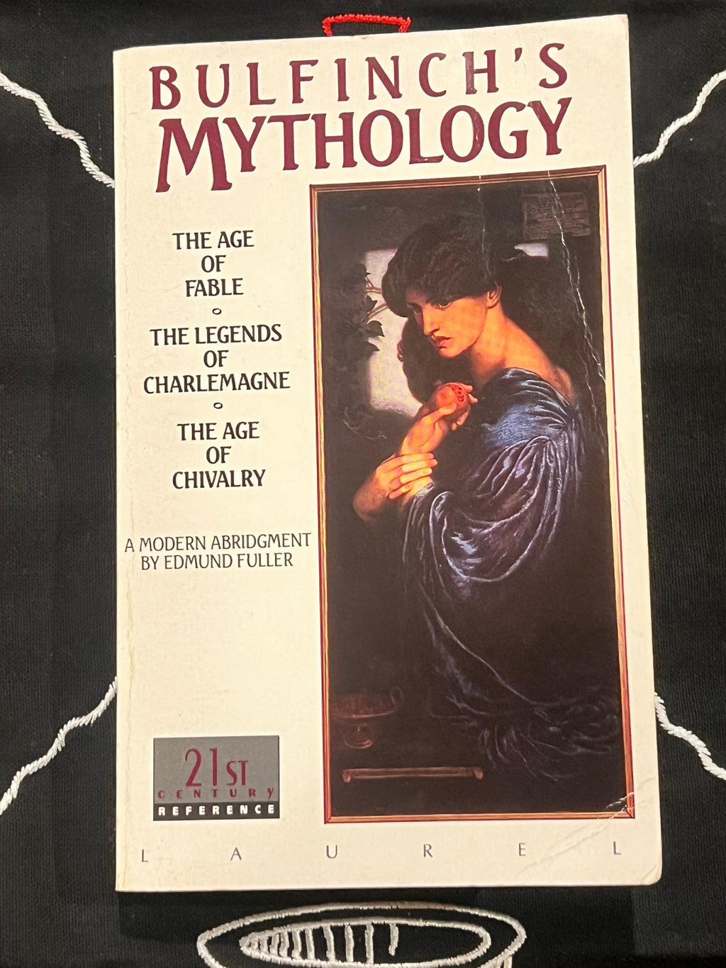 A paperback book of 'Bulfinch's Mythology' by Edmund Fuller, published by Laurel, displayed on a black fabric background with white embroidered patterns.