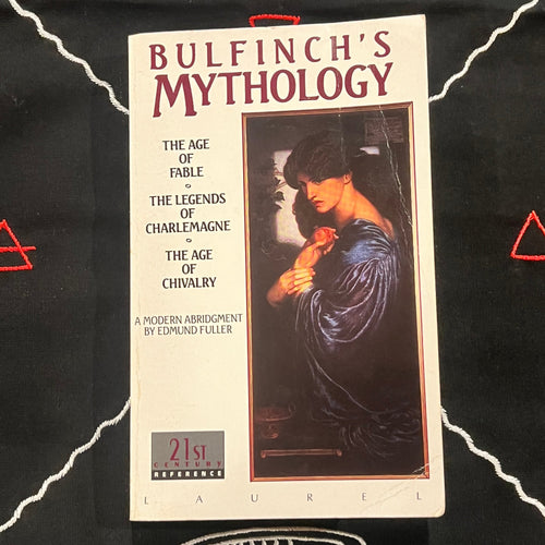 A paperback book of 'Bulfinch's Mythology' by Edmund Fuller, published by Laurel, displayed on a black fabric background with white embroidered patterns.