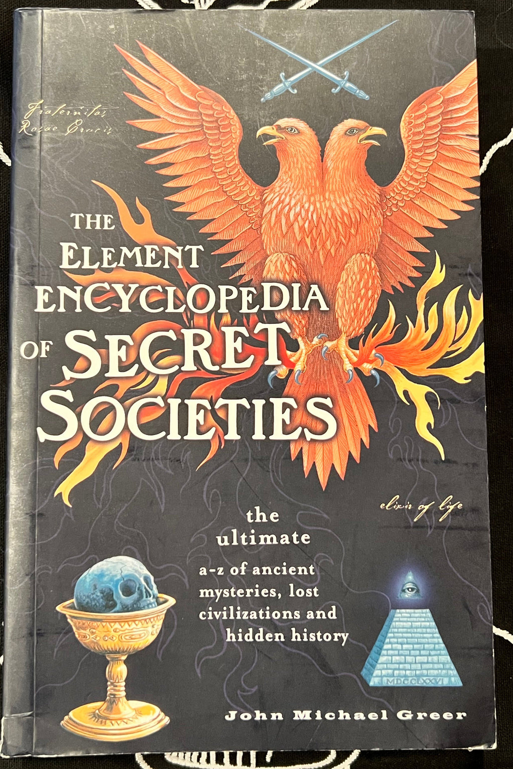 The Element Encyclopedia of Secret Societies book by John Michael Greer, featuring a dark cover with a large orange phoenix, crossed swords, a blue skull in a gold cup, and a blue pyramid.