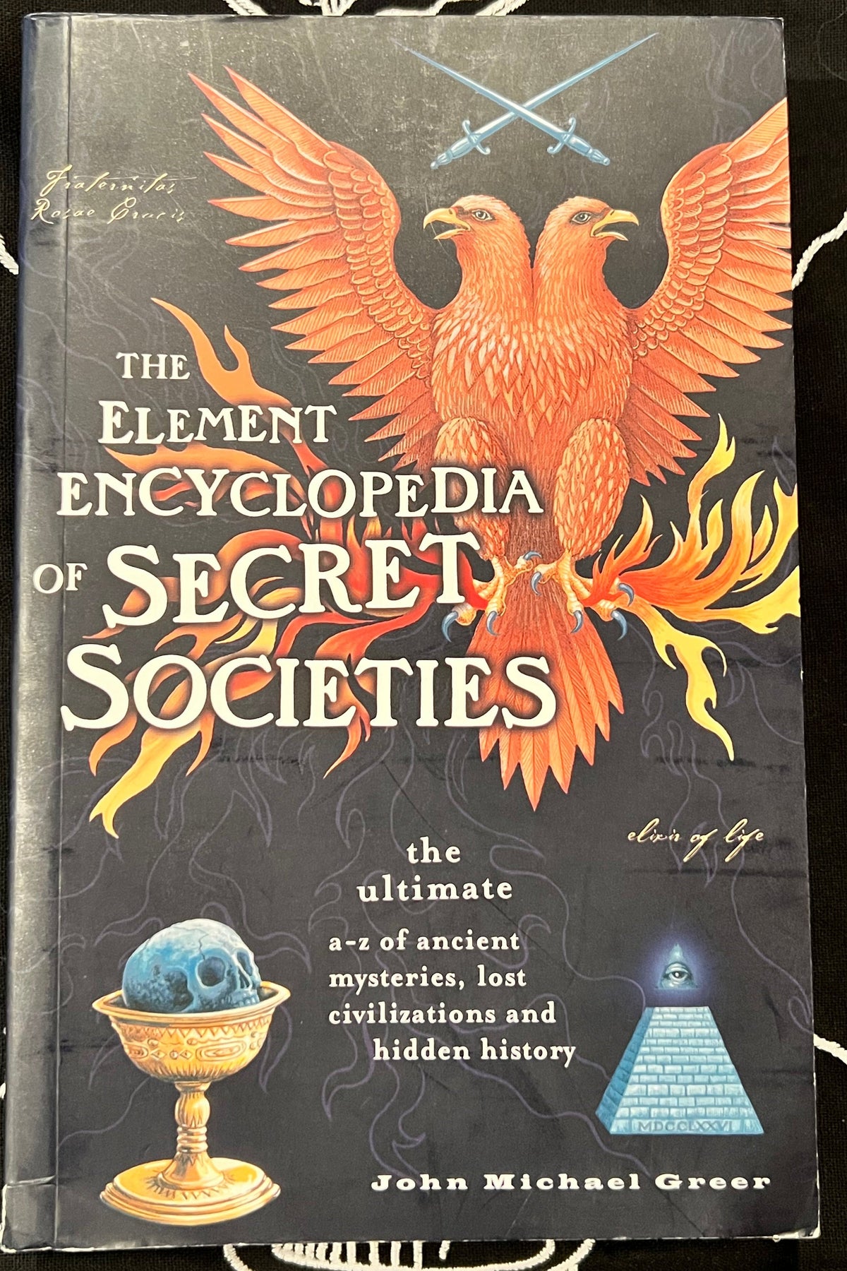 The Element Encyclopedia of Secret Societies book by John Michael Greer, featuring a dark cover with a large orange phoenix, crossed swords, a blue skull in a gold cup, and a blue pyramid.