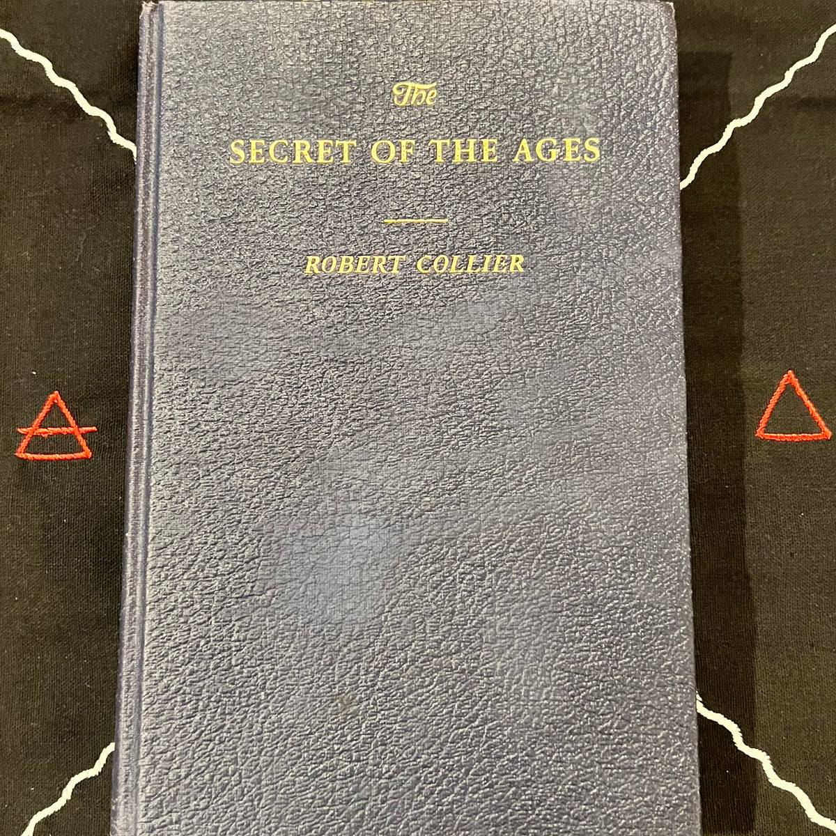 A vintage hardcover book titled 'The Secret of the Ages' by Robert Collier, featuring a textured dark blue-gray cover with gold lettering, displayed on a black background with white line art and red geometric symbols.