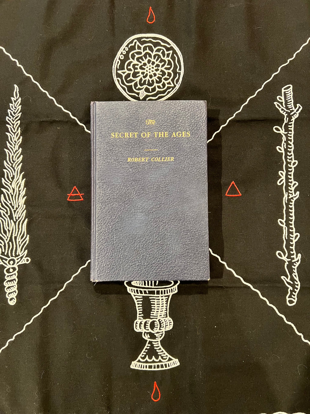 A hardcover book titled 'The Secret of the Ages' by Robert Collier, featuring a textured dark gray cover with gold lettering, displayed on a black background decorated with white occult-style illustrations and red symbols.