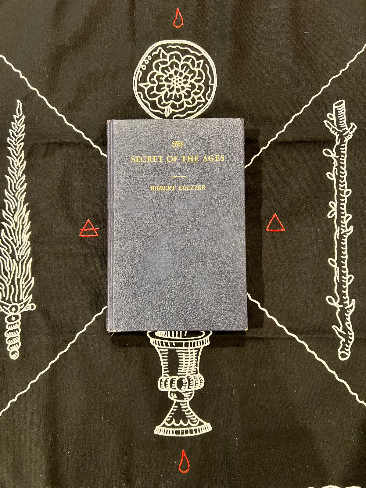 A hardcover book titled 'The Secret of the Ages' by Robert Collier, featuring a textured dark gray cover with gold lettering, displayed on a black background decorated with white occult-style illustrations and red symbols.