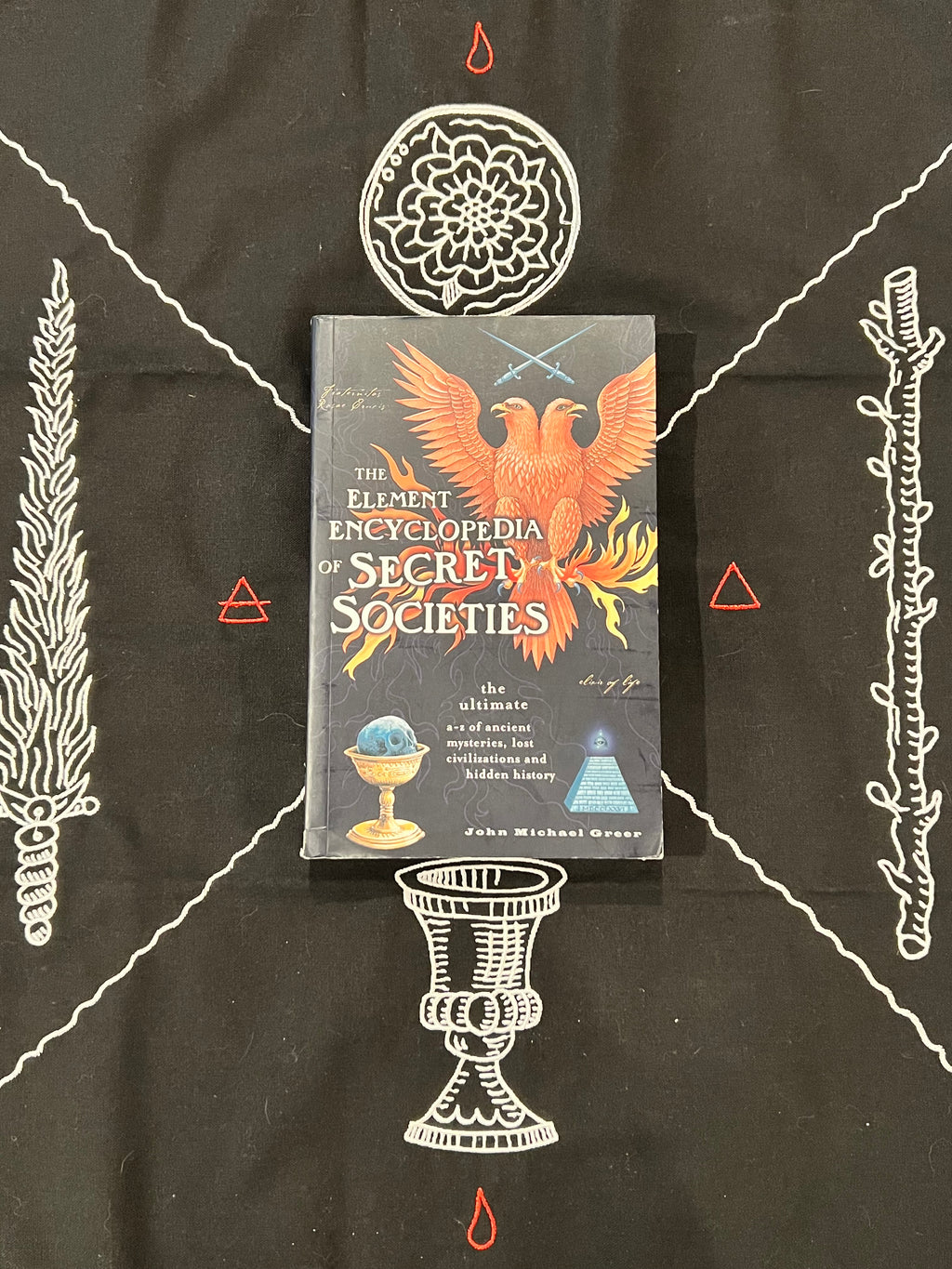 The Element Encyclopedia of Secret Societies book by John Michael Grease, featuring a dark cover with a fiery phoenix illustration, laid flat on a black fabric background decorated with white occult-style illustrations.