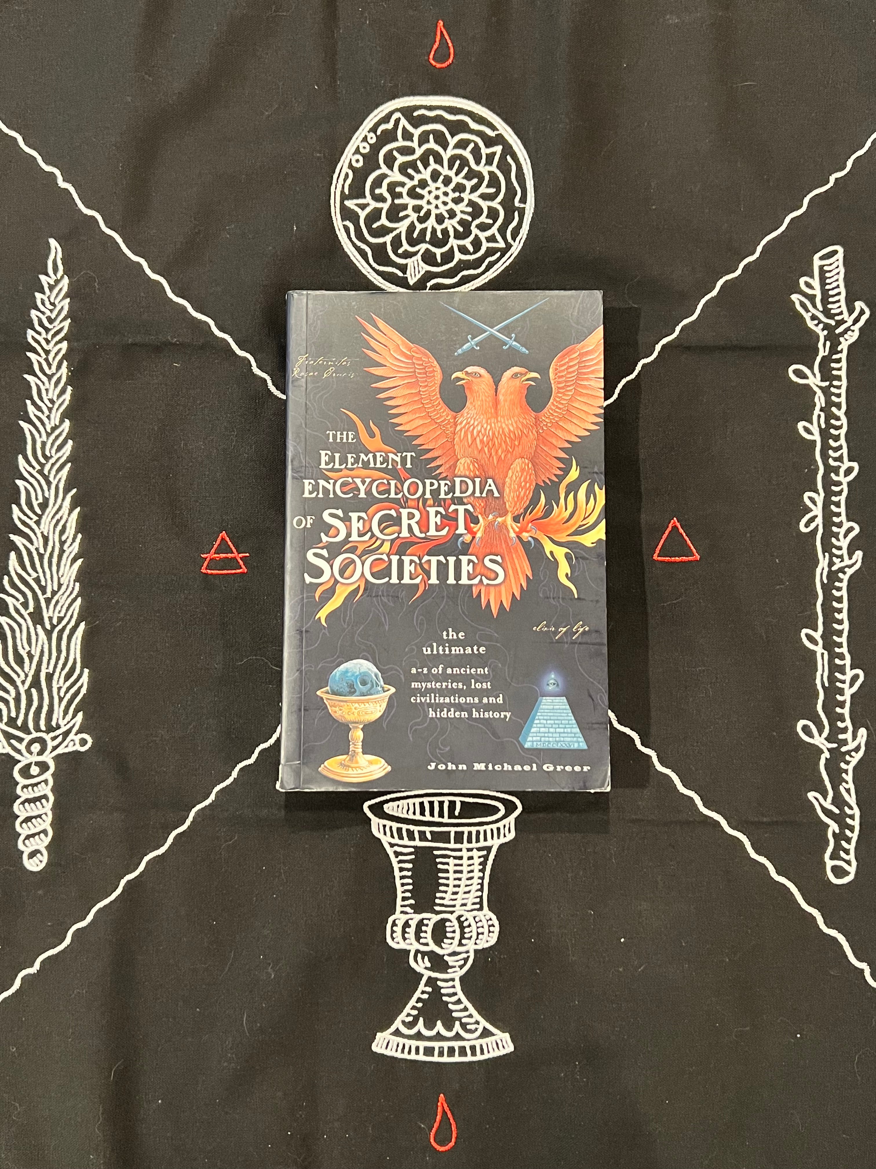 The Element Encyclopedia of Secret Societies book by John Michael Grease, featuring a dark cover with a fiery phoenix illustration, laid flat on a black fabric background decorated with white occult-style illustrations.