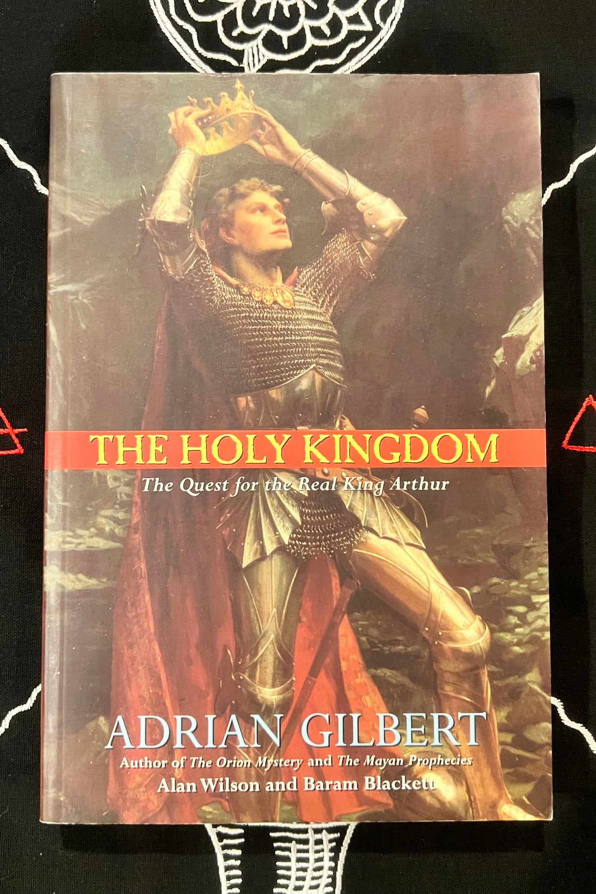 The Holy Kingdom book by Adrian Gilbert, featuring a cover illustration of a warrior in chainmail armor with a red cape and crown, set against a black background with white occult symbols.