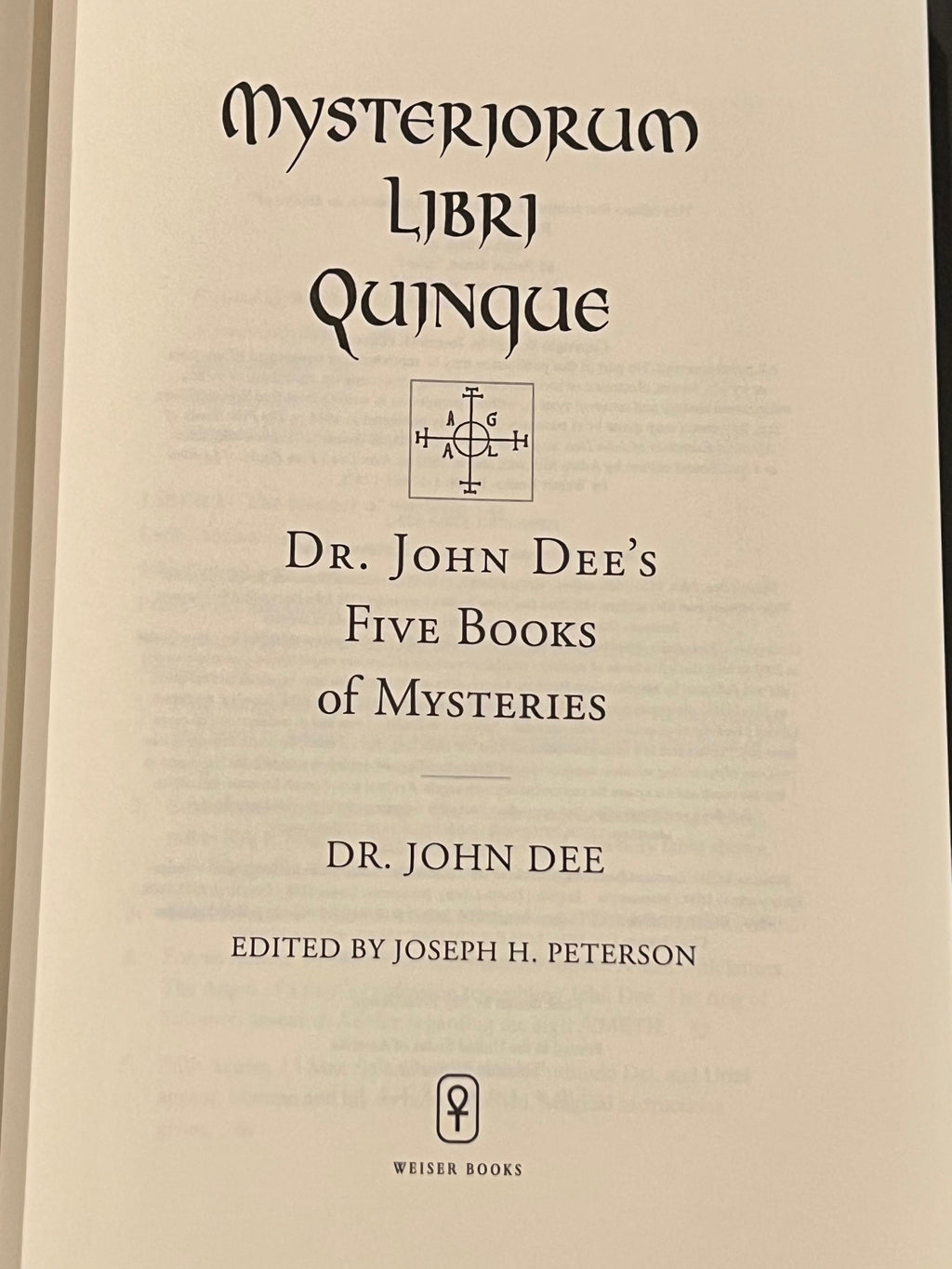 An open book showing the title page of 'Mysteriorum Libri Quinque' by Dr. John Dee, with the Weiser Books logo at the bottom, photographed against a dark patterned fabric background.