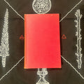 A red hardcover book lies centered on a black background printed with white occult-style illustrations and small red symbols.