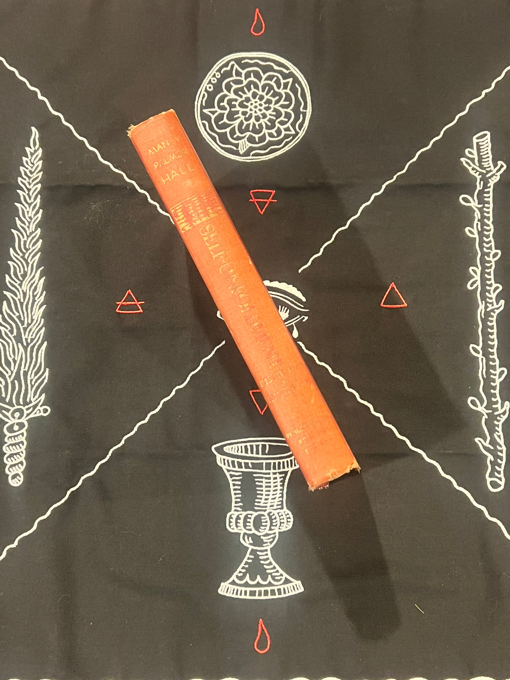 An orange book lies diagonally on a black cloth printed with white occult-style symbols, including a chalice, floral circle, and geometric lines.