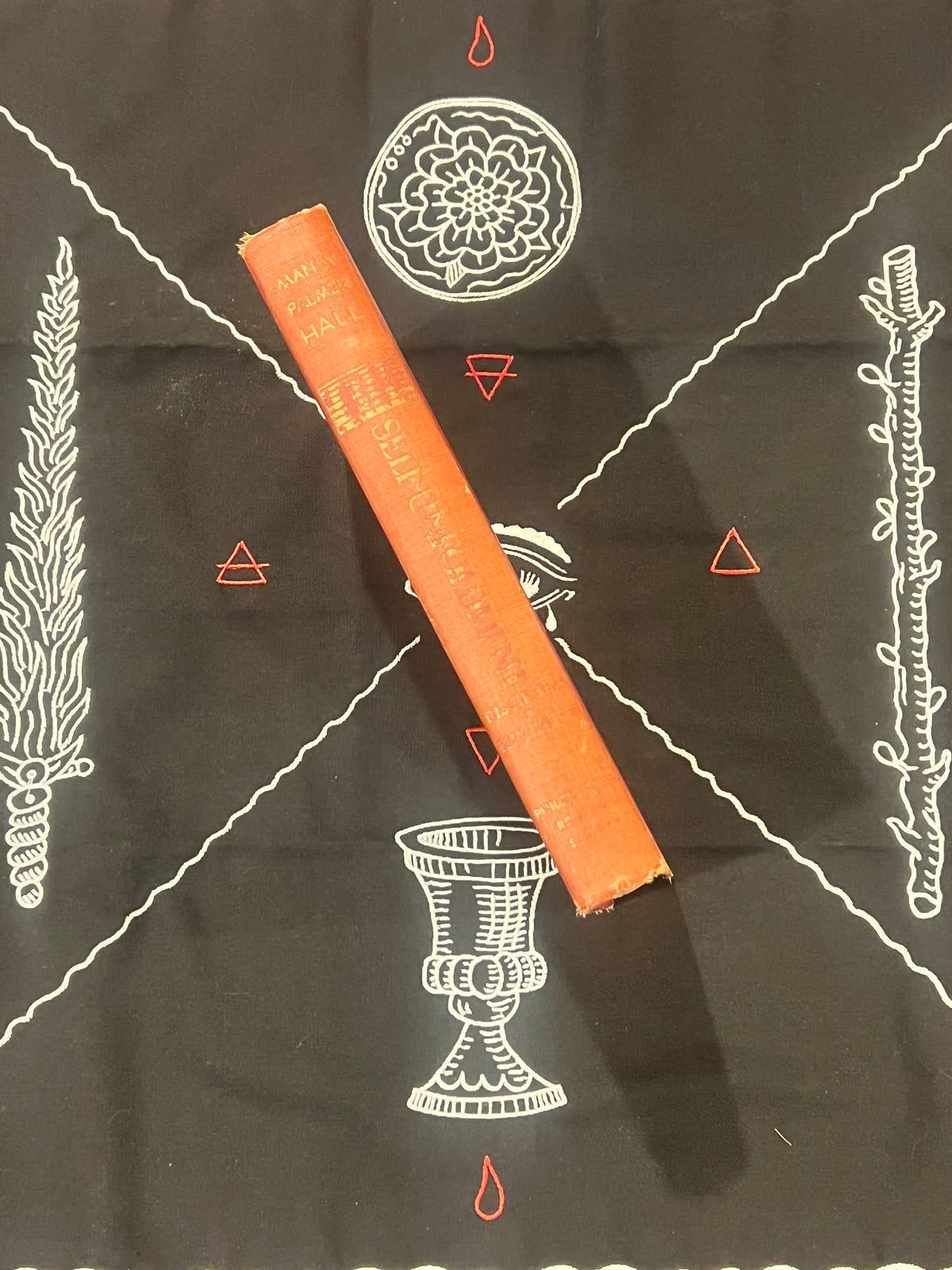 An orange book lies diagonally on a black cloth printed with white occult-style symbols, including a chalice, floral circle, and geometric lines.