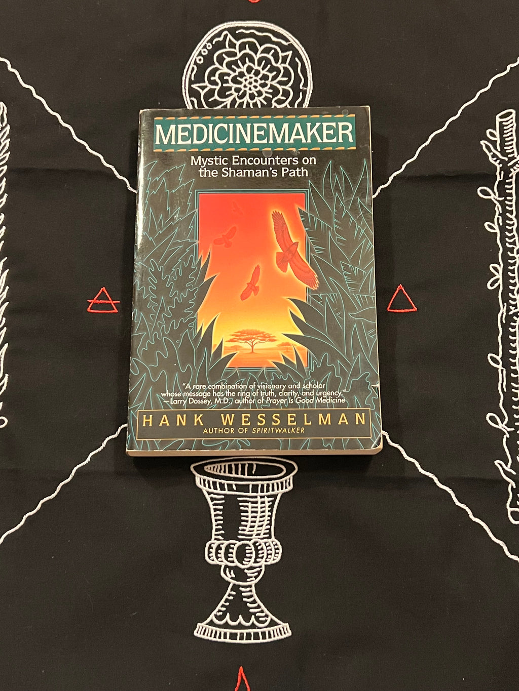 The book 'Medicinemaker' by Hank Wesselman, featuring a dark green cover with a central orange and red profile illustration, displayed on a black cloth with white and red occult-style drawings.