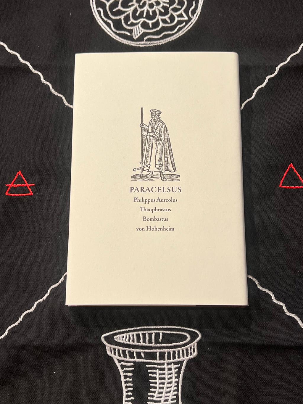 A cream-colored book titled 'PARACELSUS' by Philipp-Antonius Theophrasus Bombastus von Hohenheim, displayed flat on a black fabric background with white and red geometric patterns.