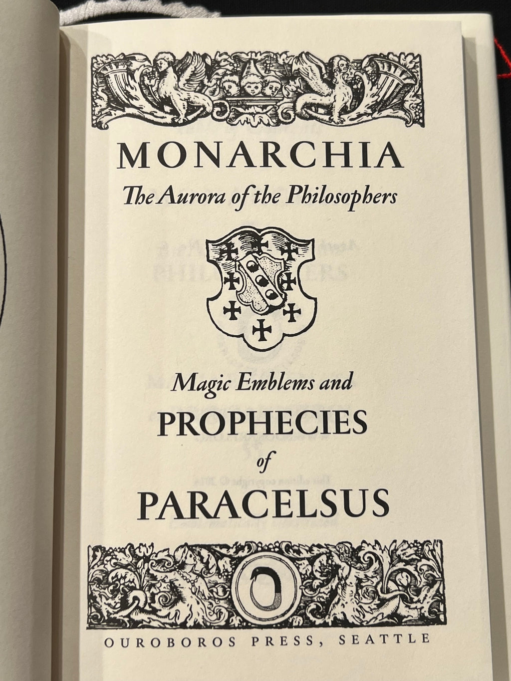 A close-up of a book's title page for 'Monarchia: The Aurora of the Philosophers' by Paracelsus, published by Ouroboros Press. The page features black gothic typography and ornate decorative borders on an off-white paper background.