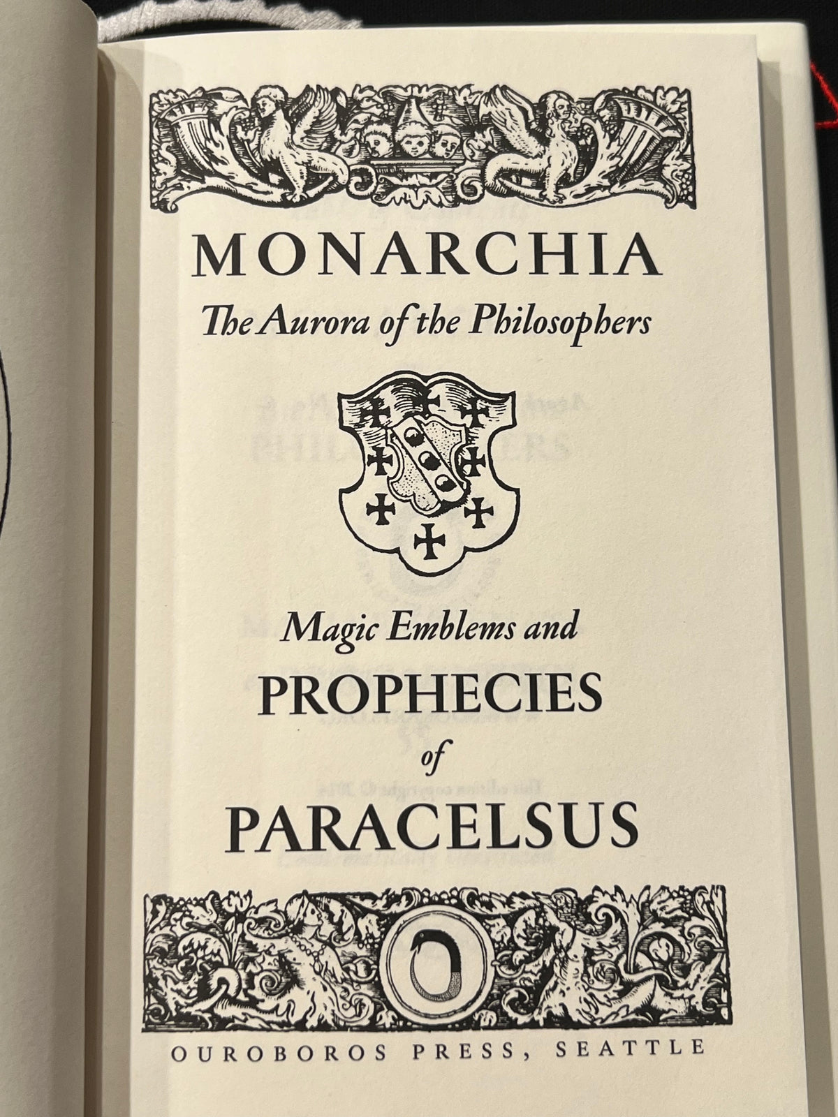A close-up of a book's title page for 'Monarchia: The Aurora of the Philosophers' by Paracelsus, published by Ouroboros Press. The page features black gothic typography and ornate decorative borders on an off-white paper background.