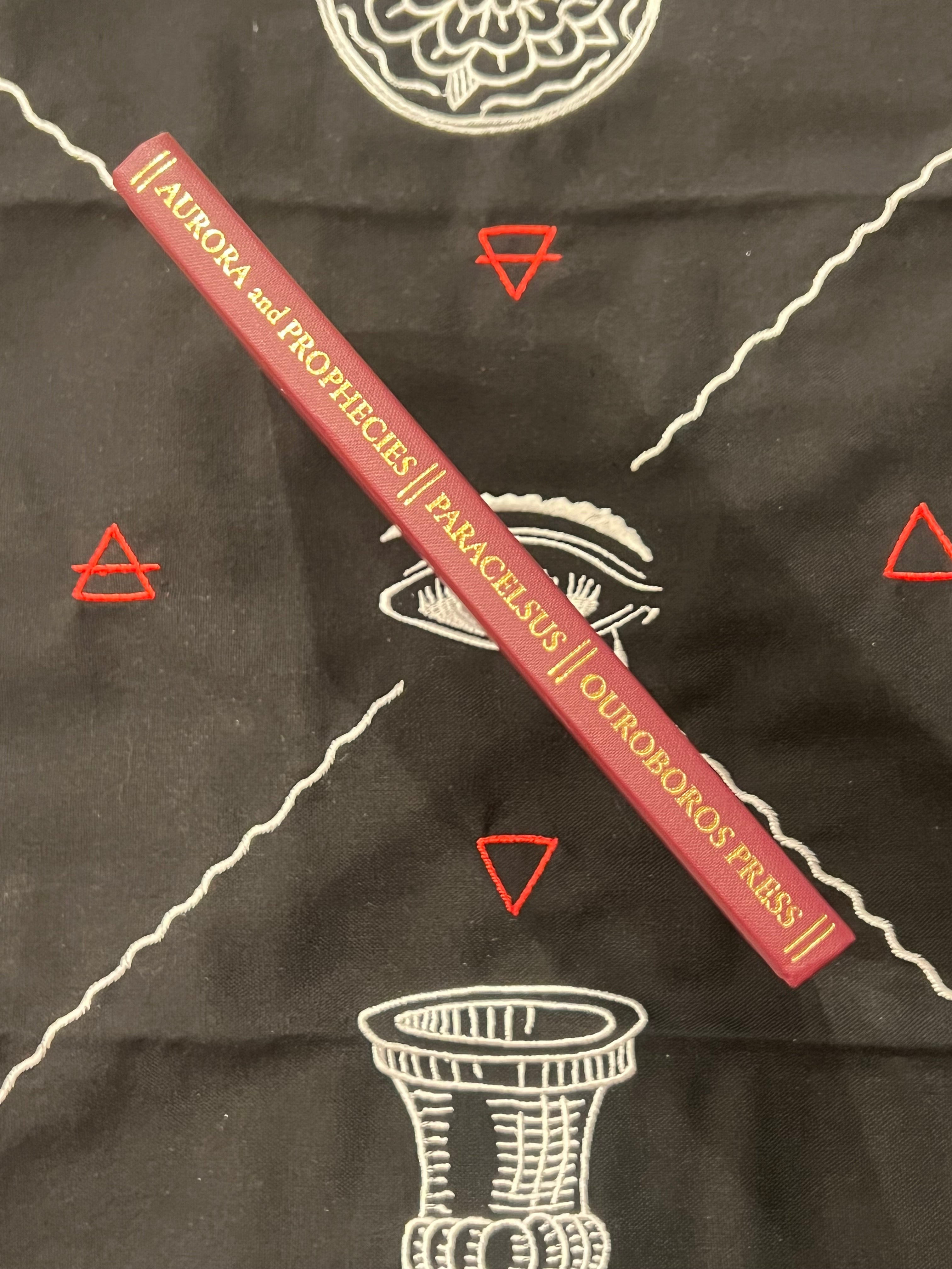 A red hardcover book titled 'Aurora and Prophecies // Paracelsus // Ouphoros Triss' lies diagonally on a black cloth with white occult-style illustrations and red triangle symbols.