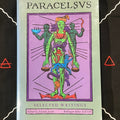 A book titled 'PARACELSVS' with 'SELECTED Writings' on the cover, featuring a colorful illustration of green and blue figures on a purple background, photographed against a black surface with white line art and red triangle symbols.