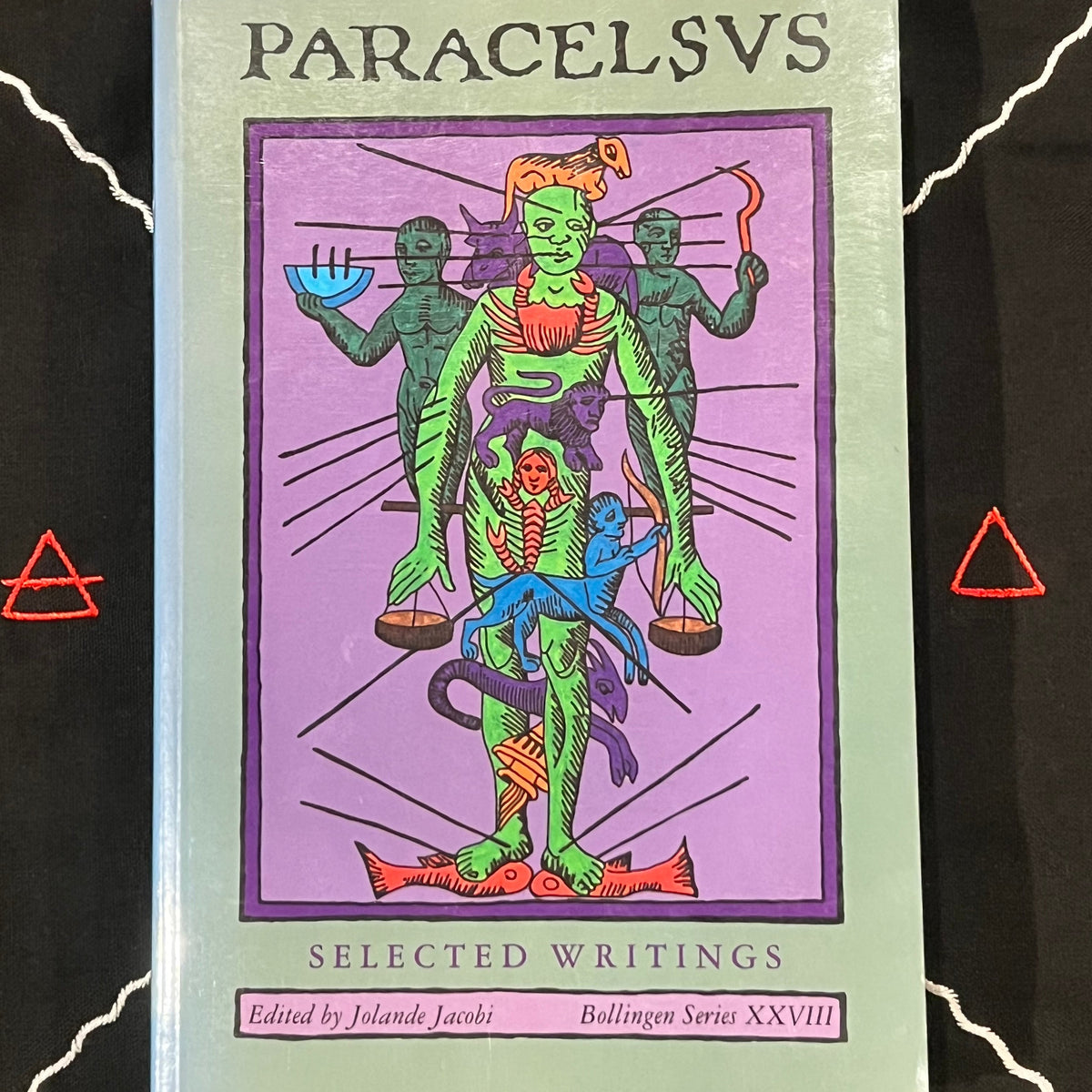 A book titled 'PARACELSVS' with 'SELECTED Writings' on the cover, featuring a colorful illustration of green and blue figures on a purple background, photographed against a black surface with white line art and red triangle symbols.