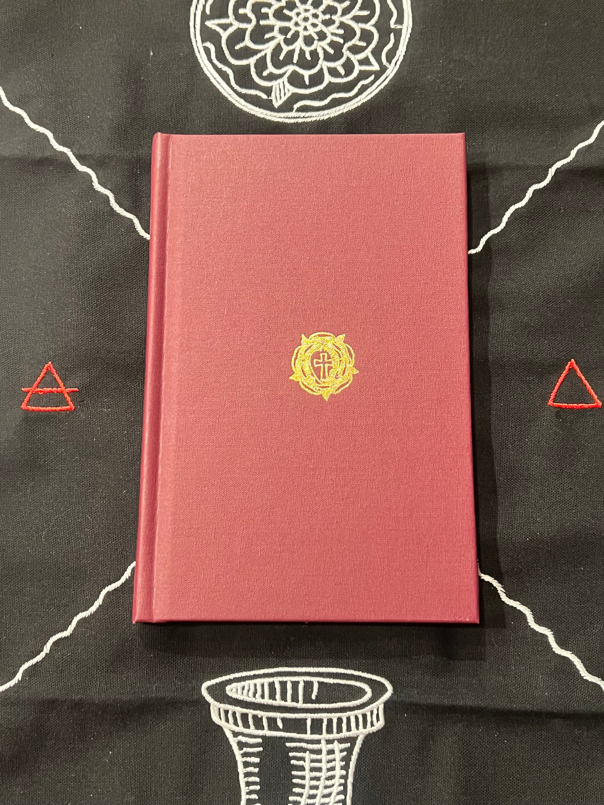 A hardcover edition of RC Manifestos in a muted brick red color, featuring a small gold circular emblem on the front cover, laid flat on a black fabric background with white and red geometric line drawings.