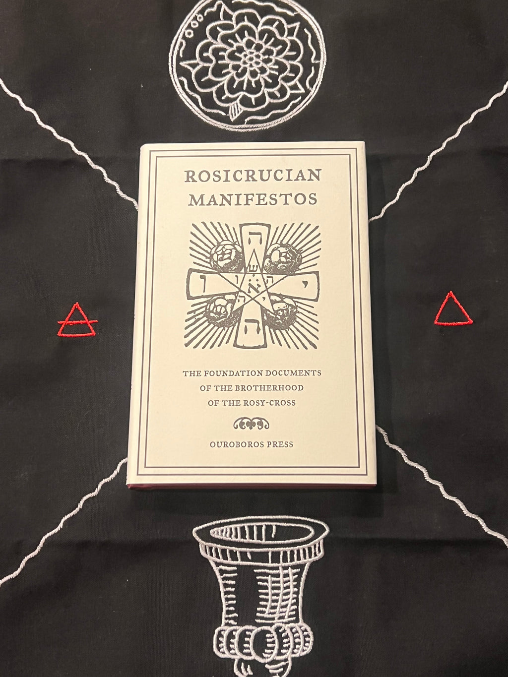 A cream-colored book titled 'Rosicrucian Manifestos' by Ourorbor Press, displayed on a black cloth with white occult-style illustrations and small red triangle accents.