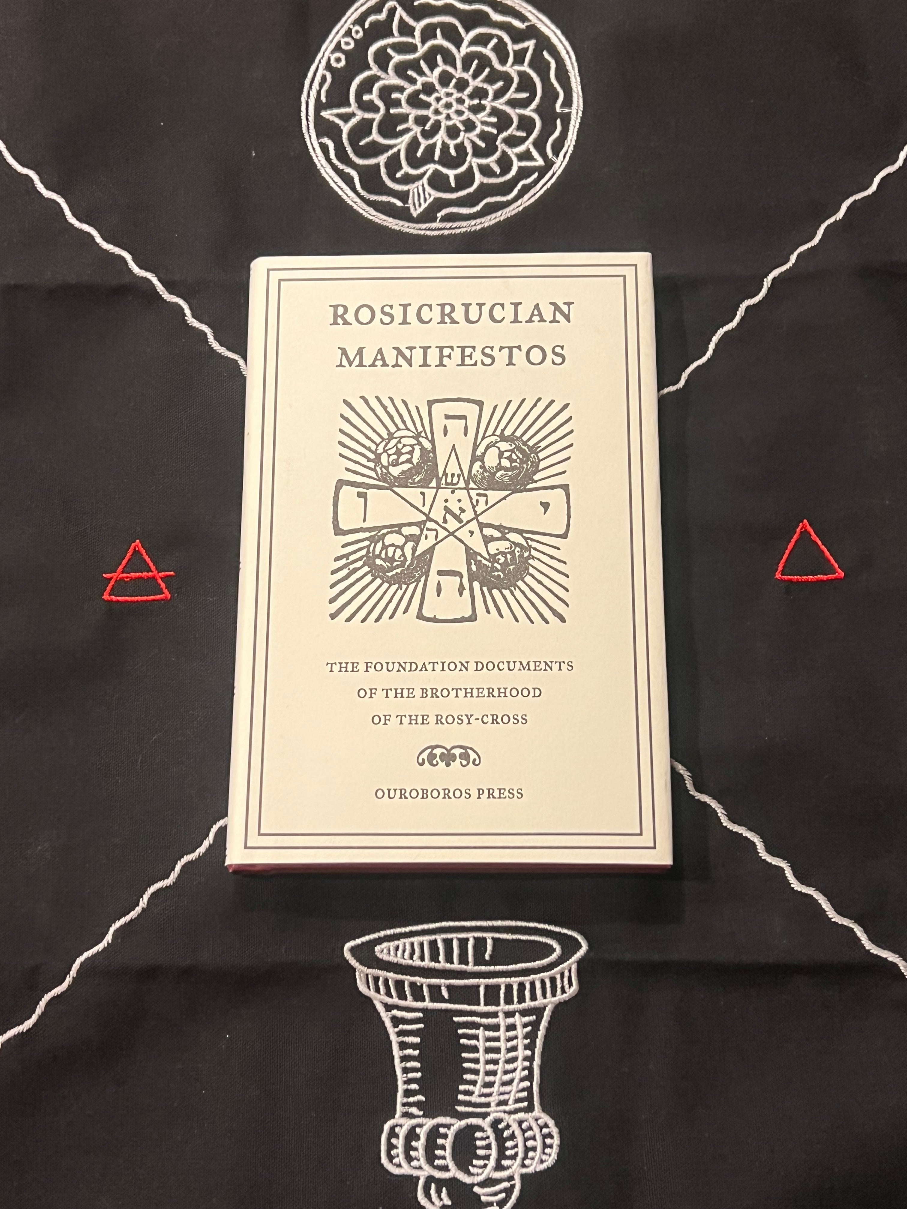 A cream-colored book titled 'Rosicrucian Manifestos' by Ourorbor Press, displayed on a black cloth with white occult-style illustrations and small red triangle accents.