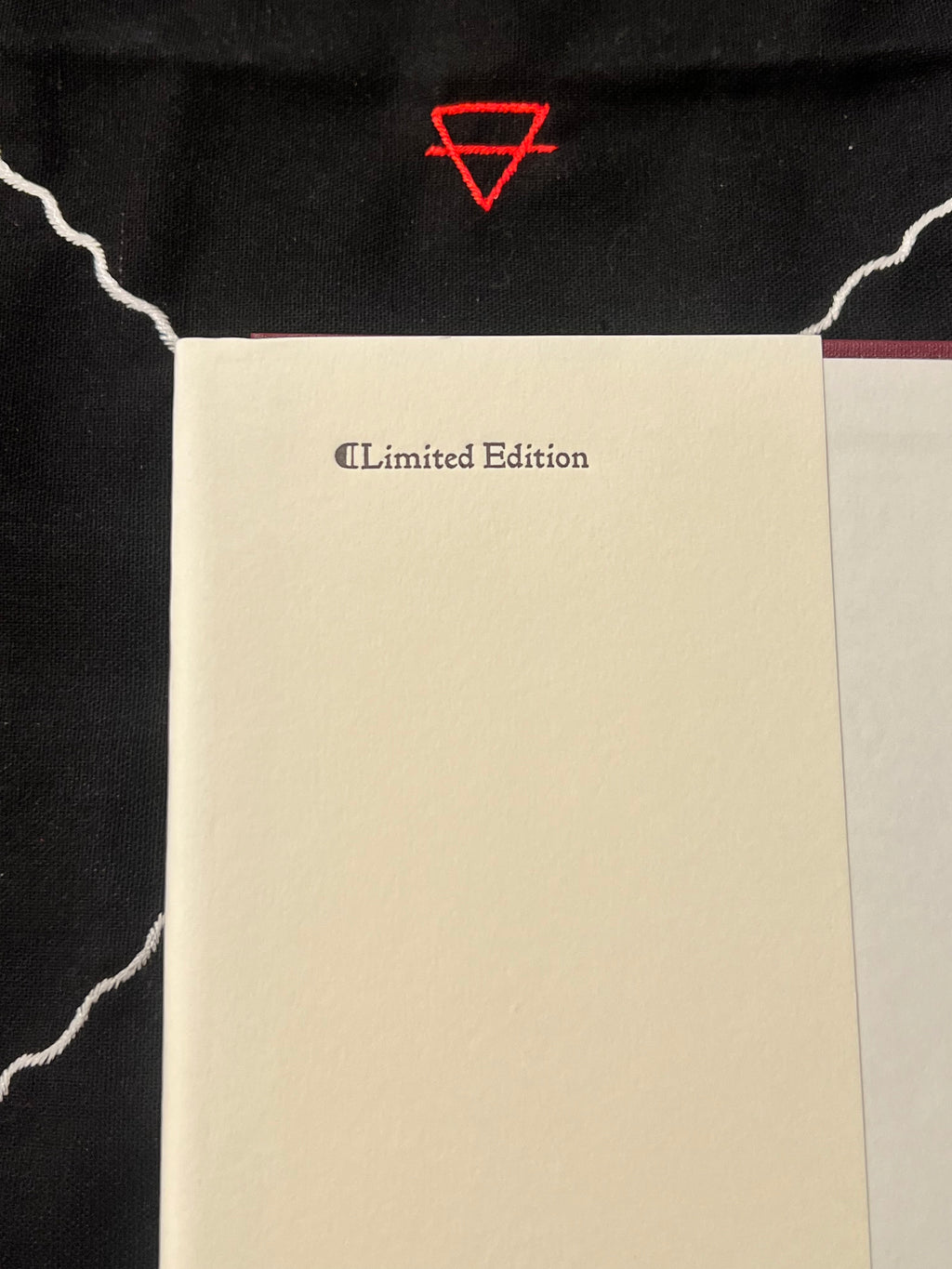 A cream-colored hardcover book or notebook with 'Limited Edition' printed in black near the top, placed on a black fabric background with a red geometric symbol above it.