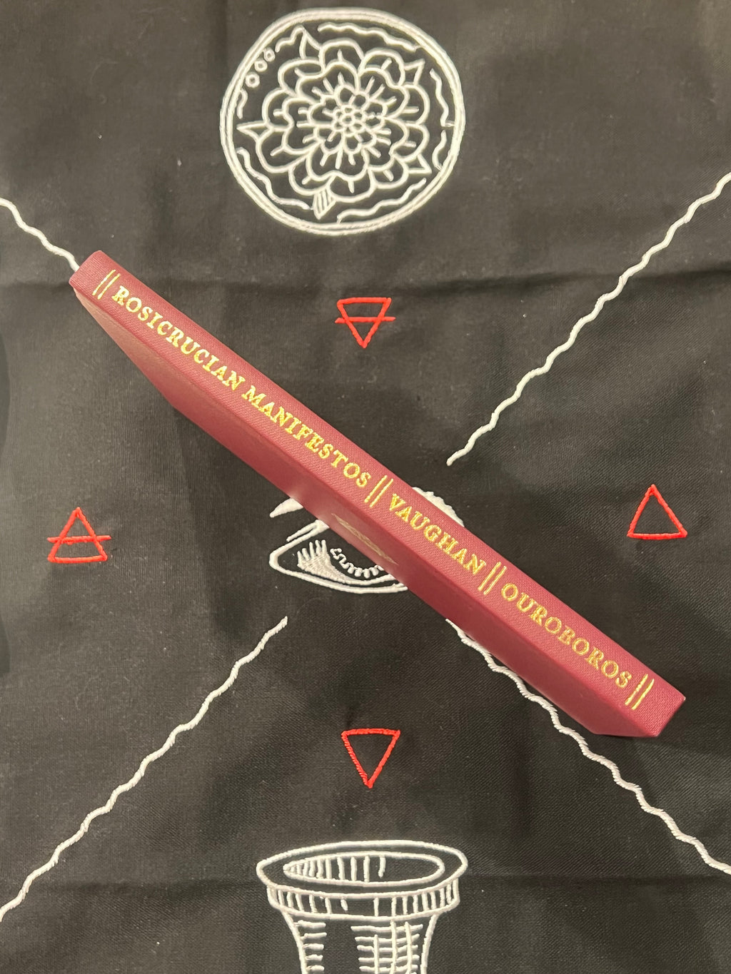 A red hardcover book titled 'Rosicrucian Manifestos // Vaughan // Ouroboros' lies diagonally on a black cloth printed with white occult-style symbols and red geometric triangles.