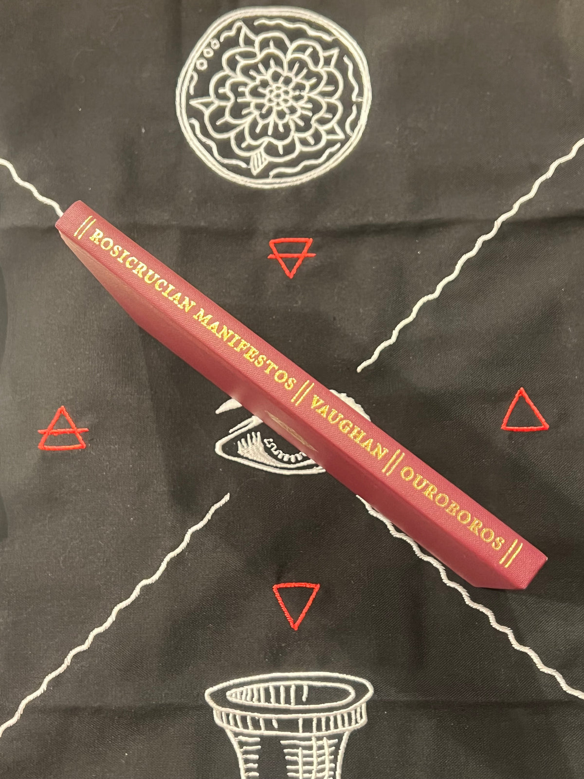 A red hardcover book titled 'Rosicrucian Manifestos // Vaughan // Ouroboros' lies diagonally on a black cloth printed with white occult-style symbols and red geometric triangles.