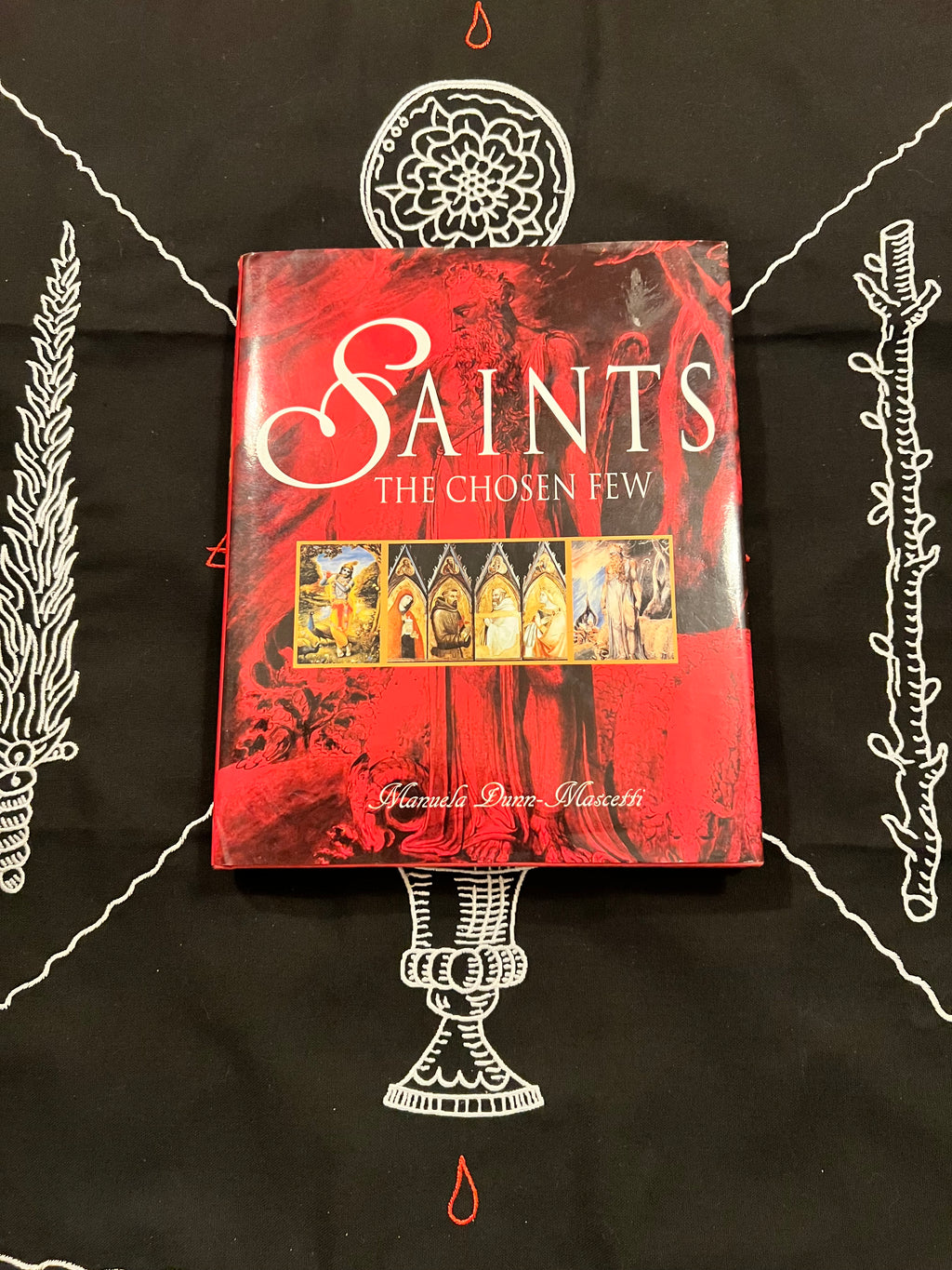 A book titled 'Saints: The Chosen Few' by Shane Peter Malicetti, featuring a red and black cover with white gothic lettering and a central illustration of saints, displayed on a black cloth with white occult-style drawings.