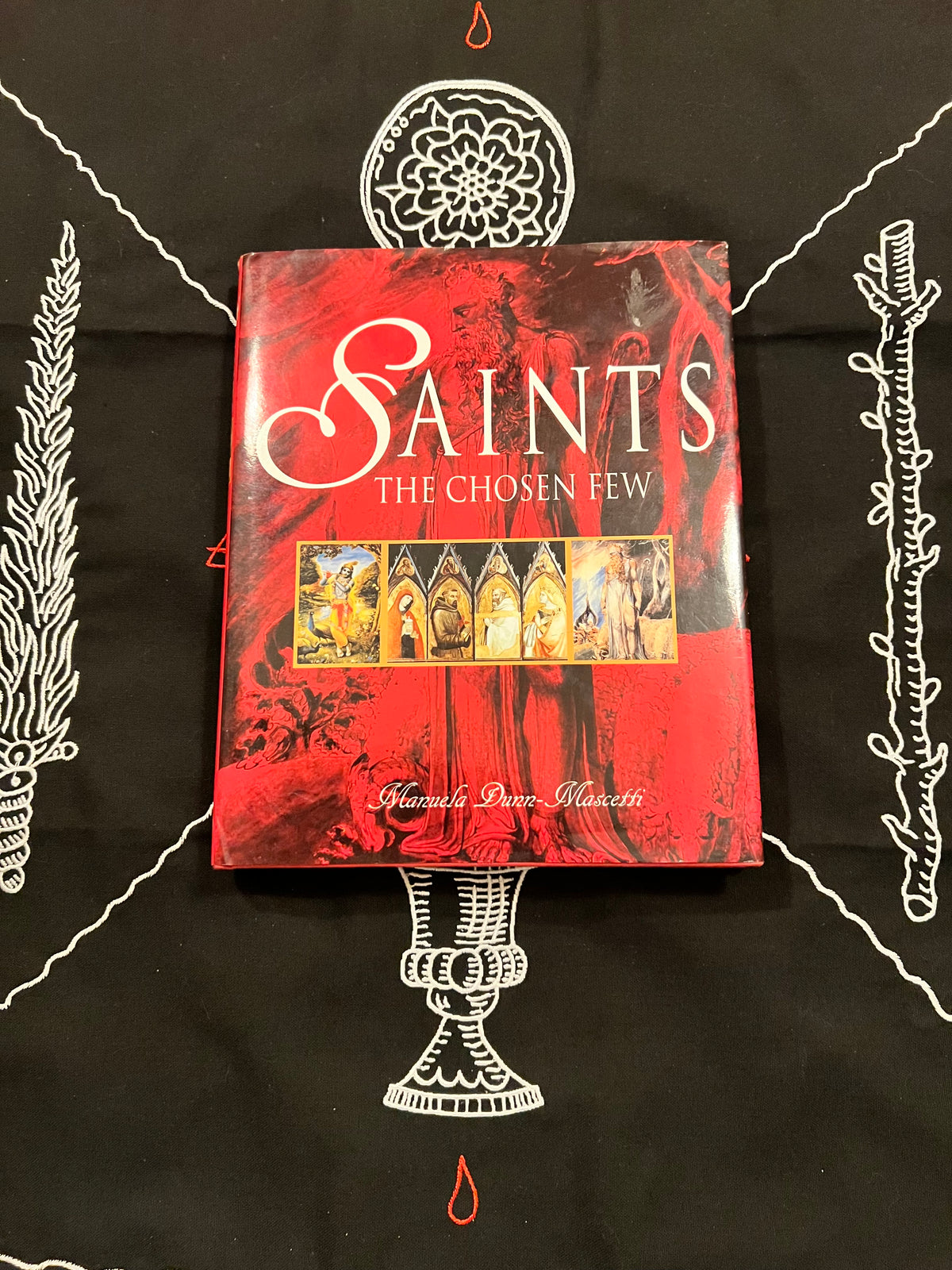 A book titled 'Saints: The Chosen Few' by Shane Peter Malicetti, featuring a red and black cover with white gothic lettering and a central illustration of saints, displayed on a black cloth with white occult-style drawings.