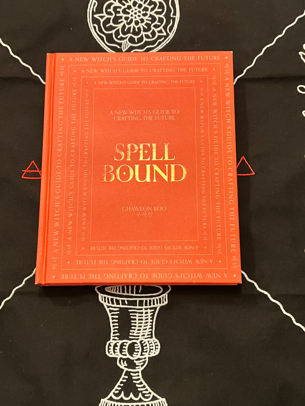 A hardcover book titled 'Spell Bound' by Chawton Roo Nae, featuring a bright orange-red cover with gold lettering, placed on a black cloth with white occult-style illustrations.