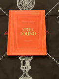 A hardcover book titled 'Spell Bound' by Chawton Roo Nae, featuring a bright orange-red cover with gold lettering, placed on a black cloth with white occult-style illustrations.