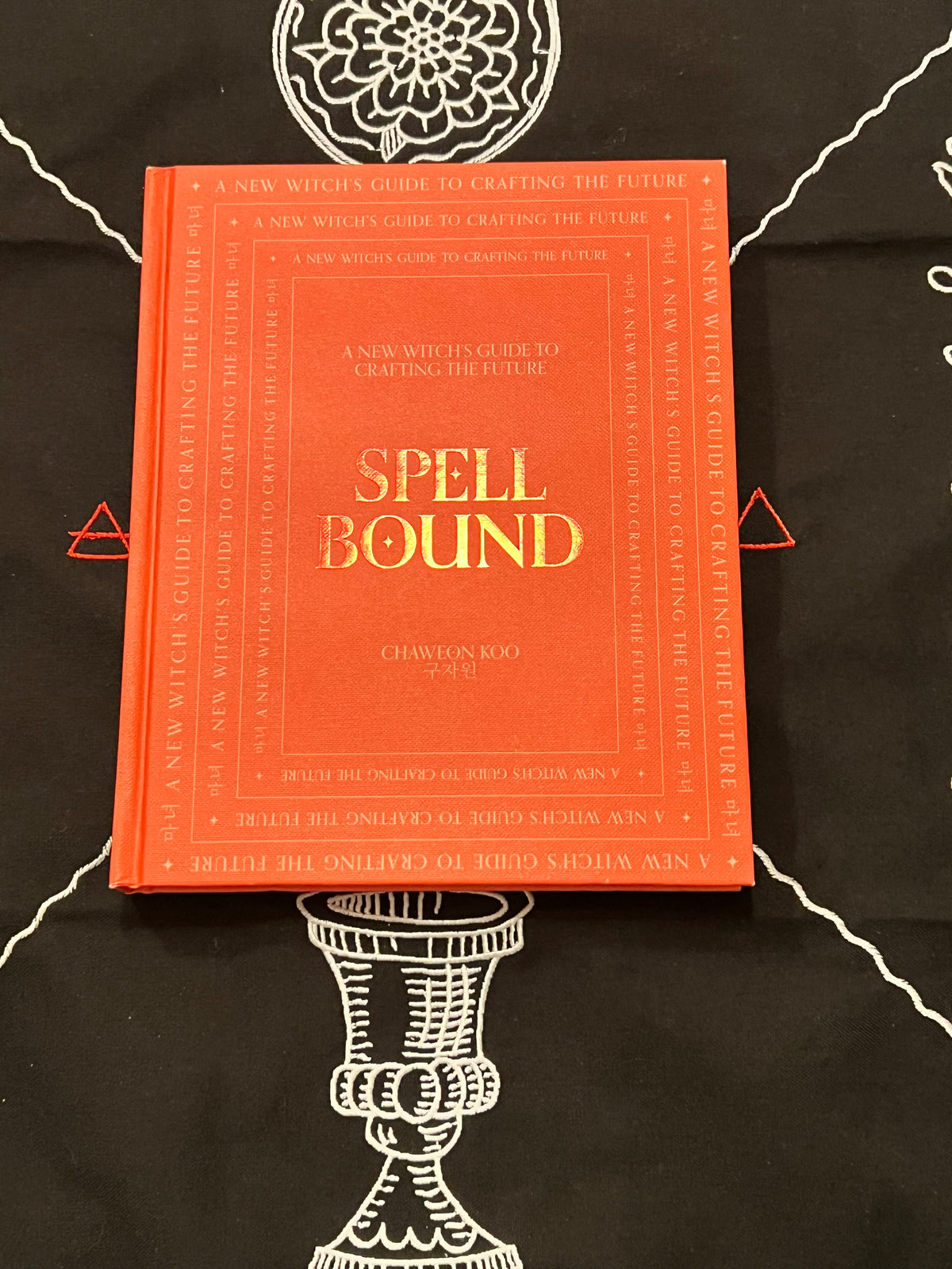 A hardcover book titled 'Spell Bound' by Chawton Roo Nae, featuring a bright orange-red cover with gold lettering, placed on a black cloth with white occult-style illustrations.
