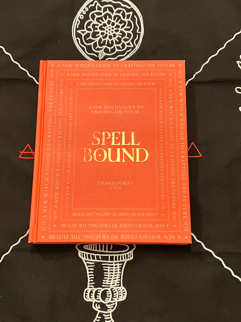 A hardcover book titled 'Spell Bound' by Charon McCoy, featuring a bright orange-red cover with gold lettering, displayed on a black background with white occult-style line drawings.