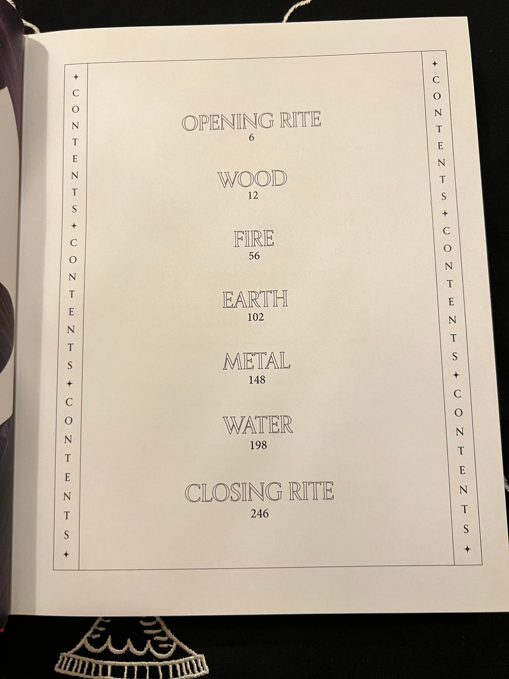 An open book or booklet showing a printed table of contents page with headings such as Opening Rite, Wood, Fire, Earth, Metal, Water, and Closing Rite, photographed against a dark background.