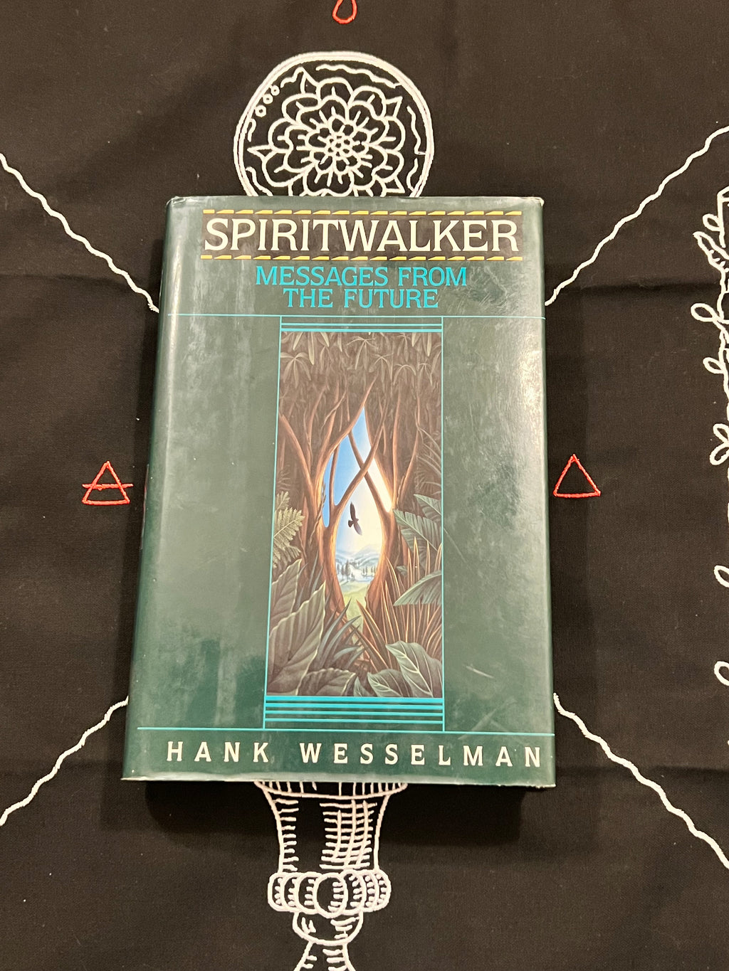The book 'Spiritwalker: Messages from the Future' by Hank Wesselman, featuring a teal-green cover with an illustration, displayed on a black cloth with white occult-style drawings.
