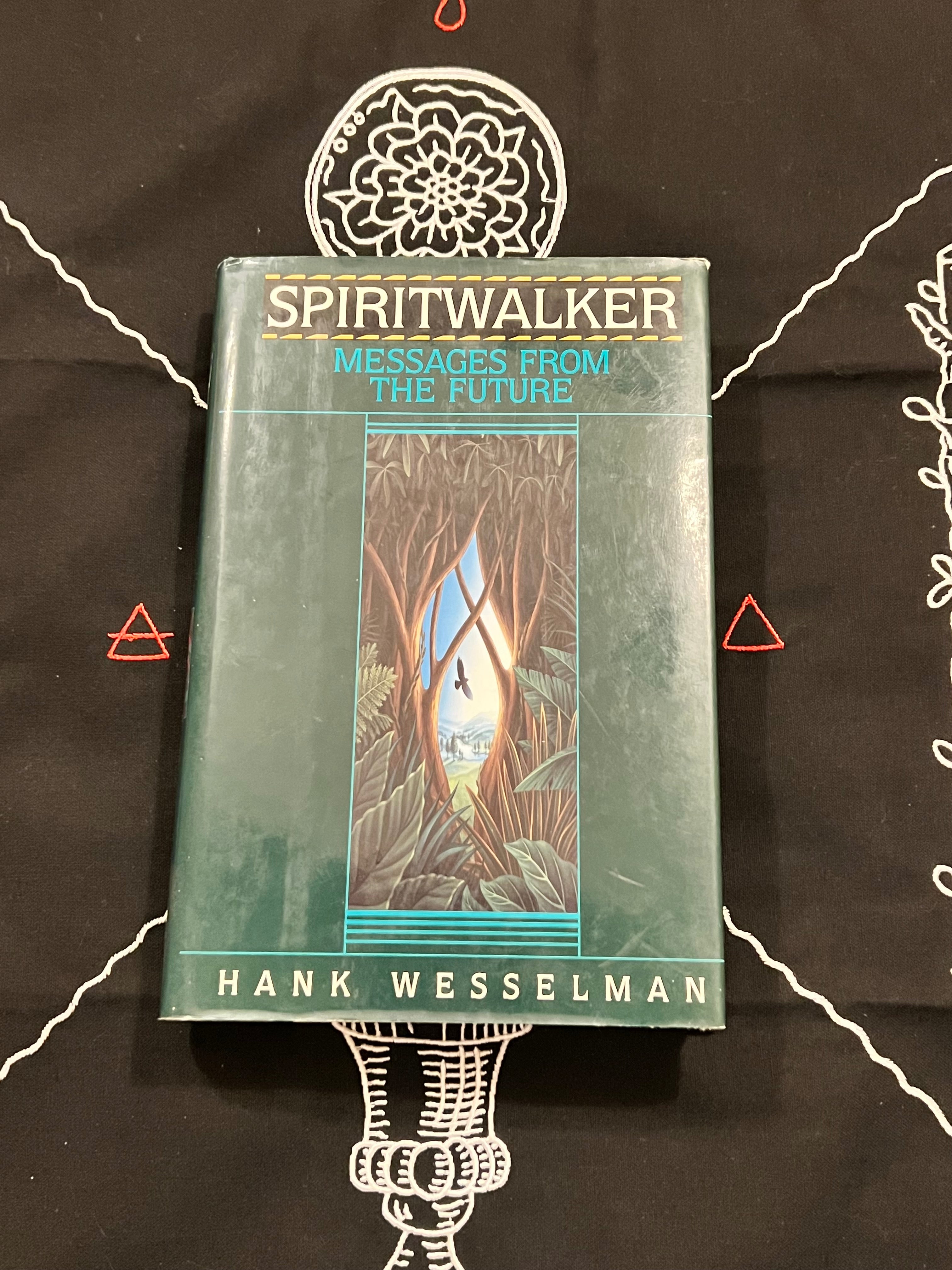 The book 'Spiritwalker: Messages from the Future' by Hank Wesselman, featuring a teal-green cover with an illustration, displayed on a black cloth with white occult-style drawings.