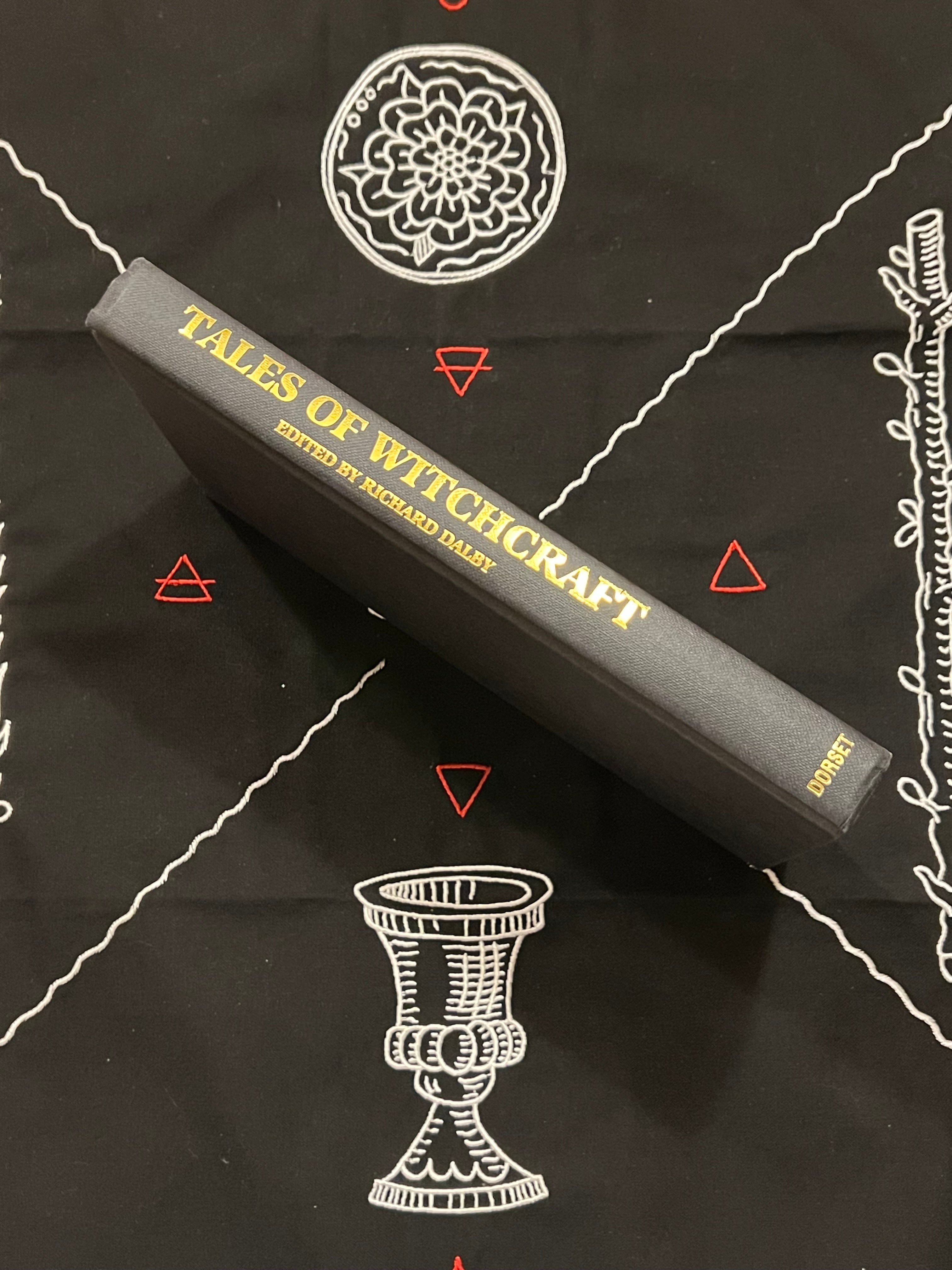 A hardcover book titled 'Tales of Witchcraft' edited by Richard Haight, lying diagonally on a black fabric background printed with white occult-style illustrations and red triangle symbols.