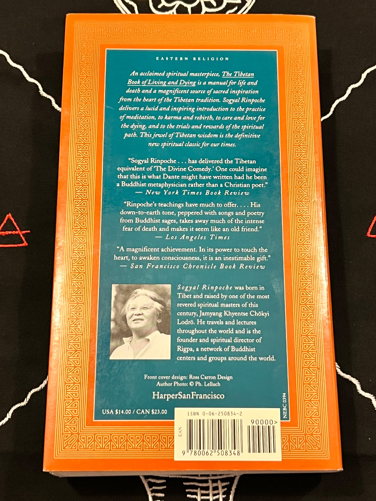 The back cover of the book 'The Tibetan Book of Living and Dying' by Sigel Rinpoche, featuring a teal blue background with white text, a small portrait photo, and an orange border, photographed flat on a black surface.