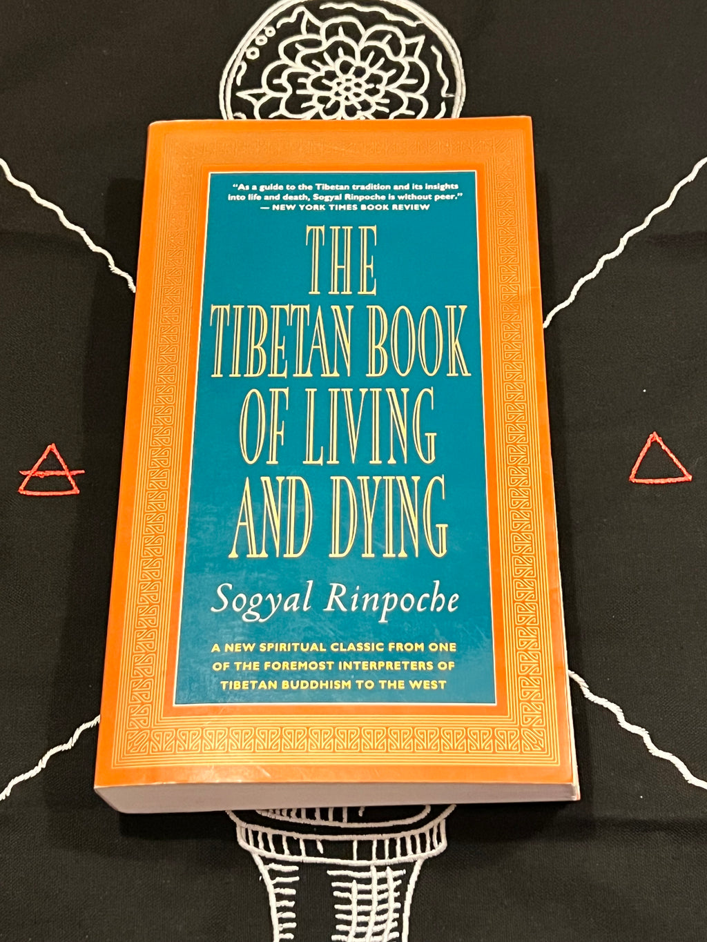The Tibetan Book of Living and Dying by Soyyal Rinpoche, featuring a bright orange cover with a teal-blue central panel and gold lettering, displayed on a black background with white and red geometric designs.