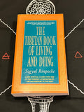 The Tibetan Book of Living and Dying by Soyyal Rinpoche, featuring a bright orange cover with a teal-blue central panel and gold lettering, displayed on a black background with white and red geometric designs.