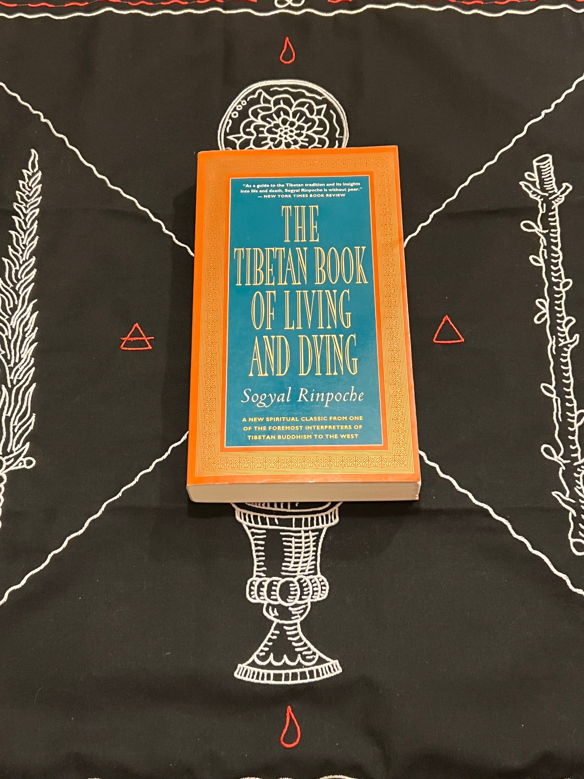 The Tibetan Book of Living and Dying by Sogyal Rinpoche, featuring an orange hardcover with a teal-blue front cover, placed on a black embroidered cloth with white and red thread details.