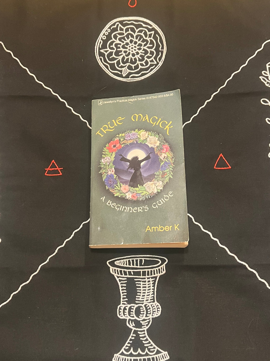 A book titled 'True Magick: A Beginner's Guide' by Amber K, placed on a black cloth printed with white occult-style diagrams and symbols.
