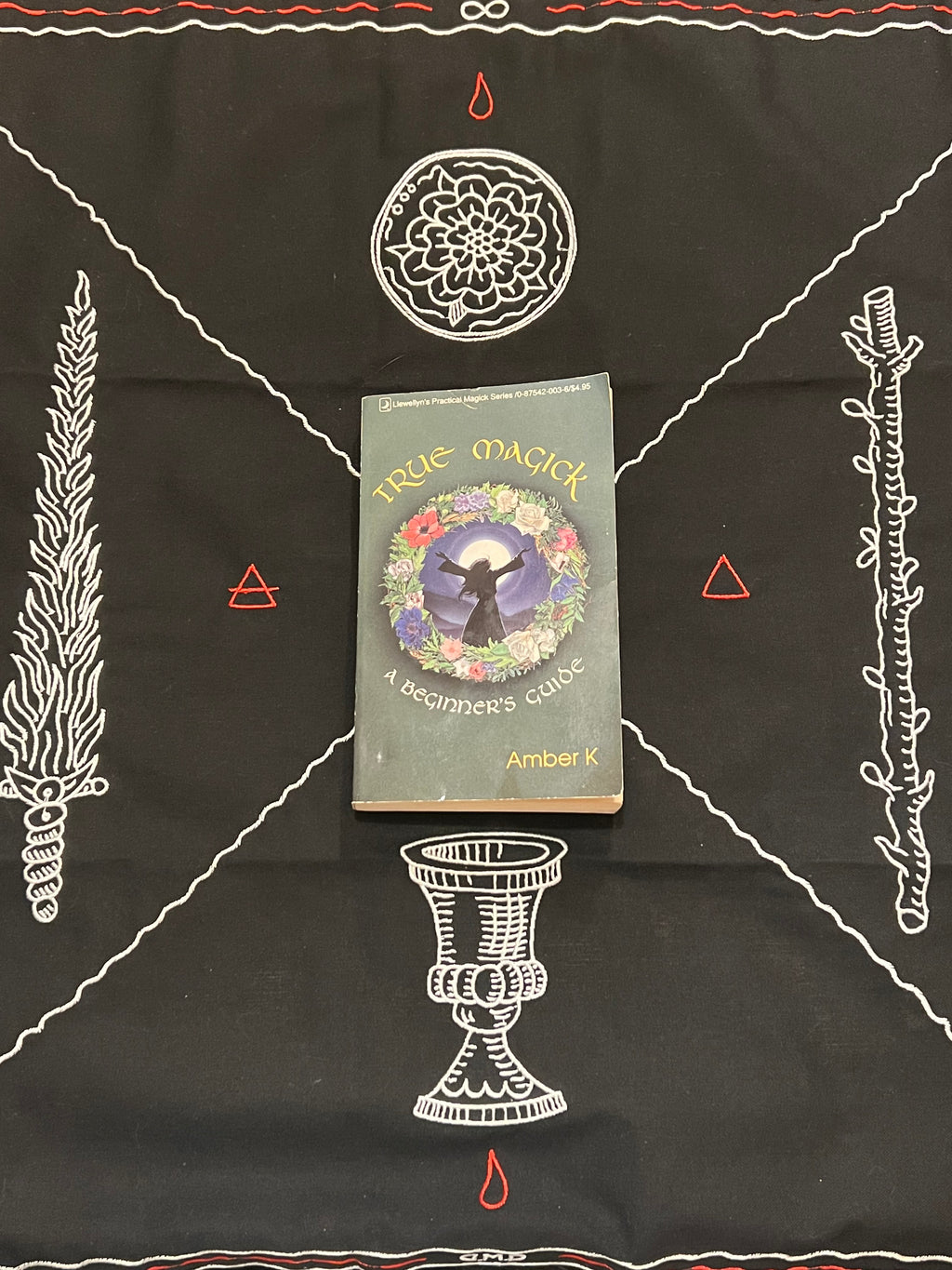 A book titled 'True Magick: A Beginner's Guide' by Amber K is placed on a black altar cloth decorated with white occult symbols, including a pentacle, wand, chalice, and triangles.