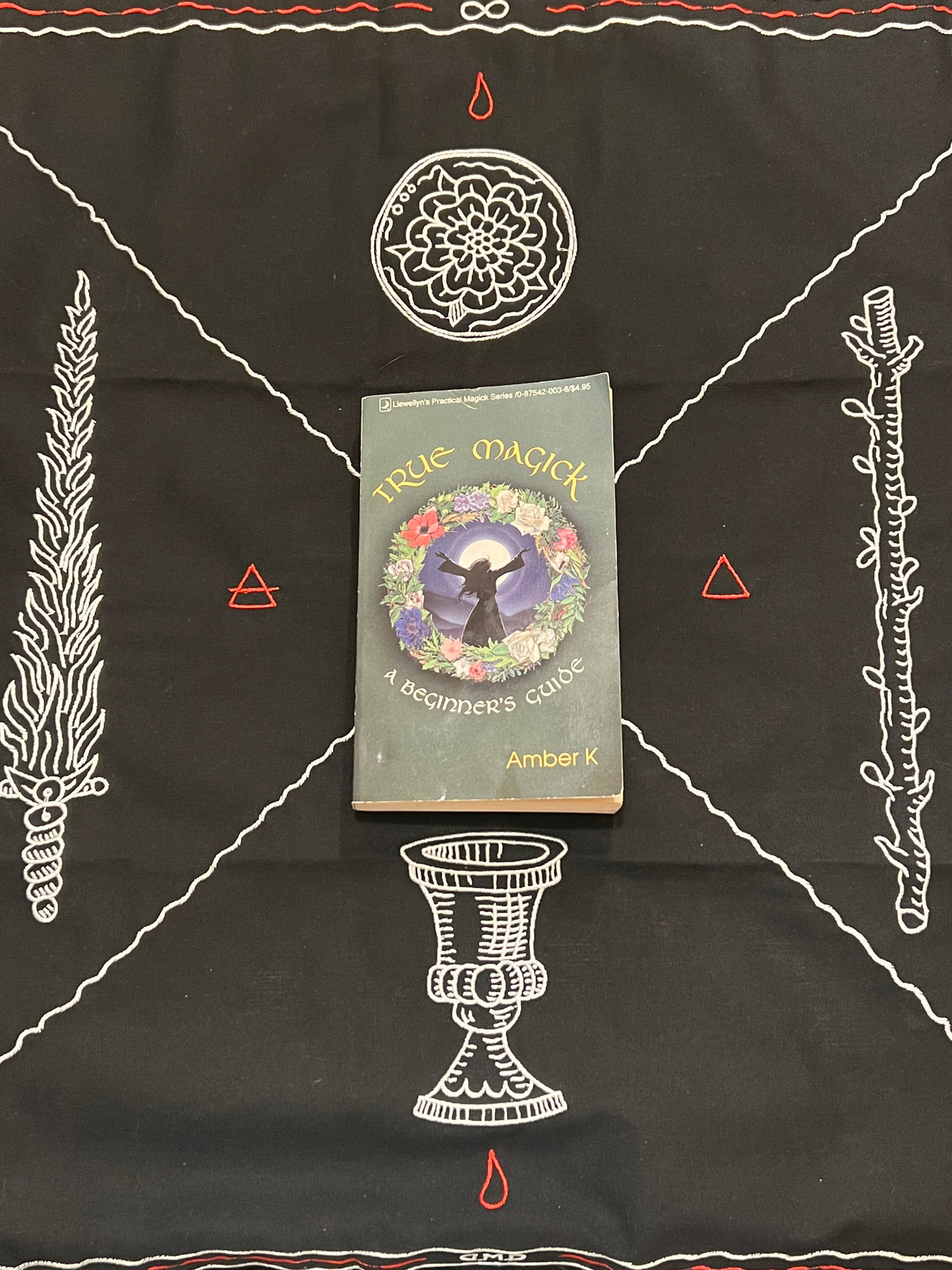 A book titled 'True Magick: A Beginner's Guide' by Amber K is placed on a black altar cloth decorated with white occult symbols, including a pentacle, wand, chalice, and triangles.